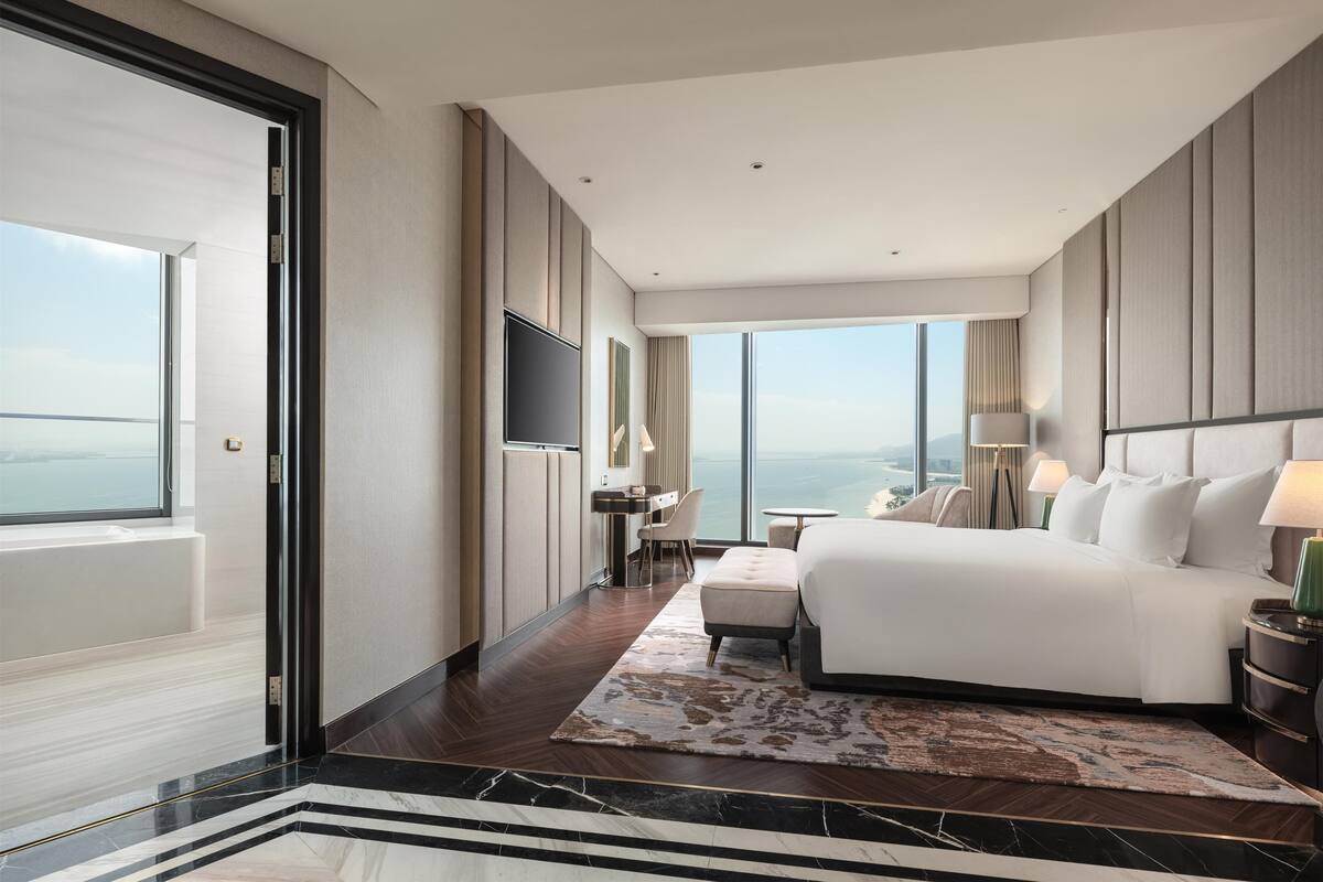 A modern hotel room with a large bed, a desk, a chair, a TV, and a view of the ocean.