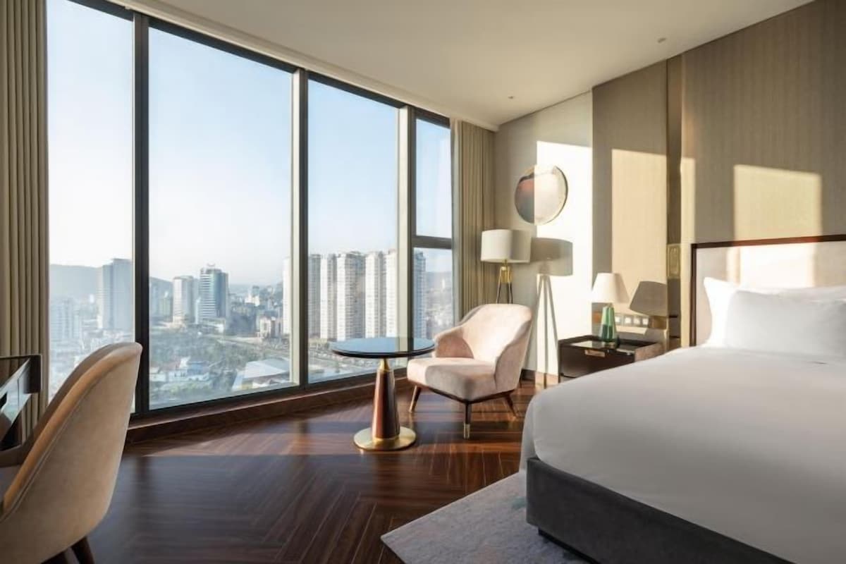 A hotel room with a large bed, a chair, a small table, and a view of the city through large windows.