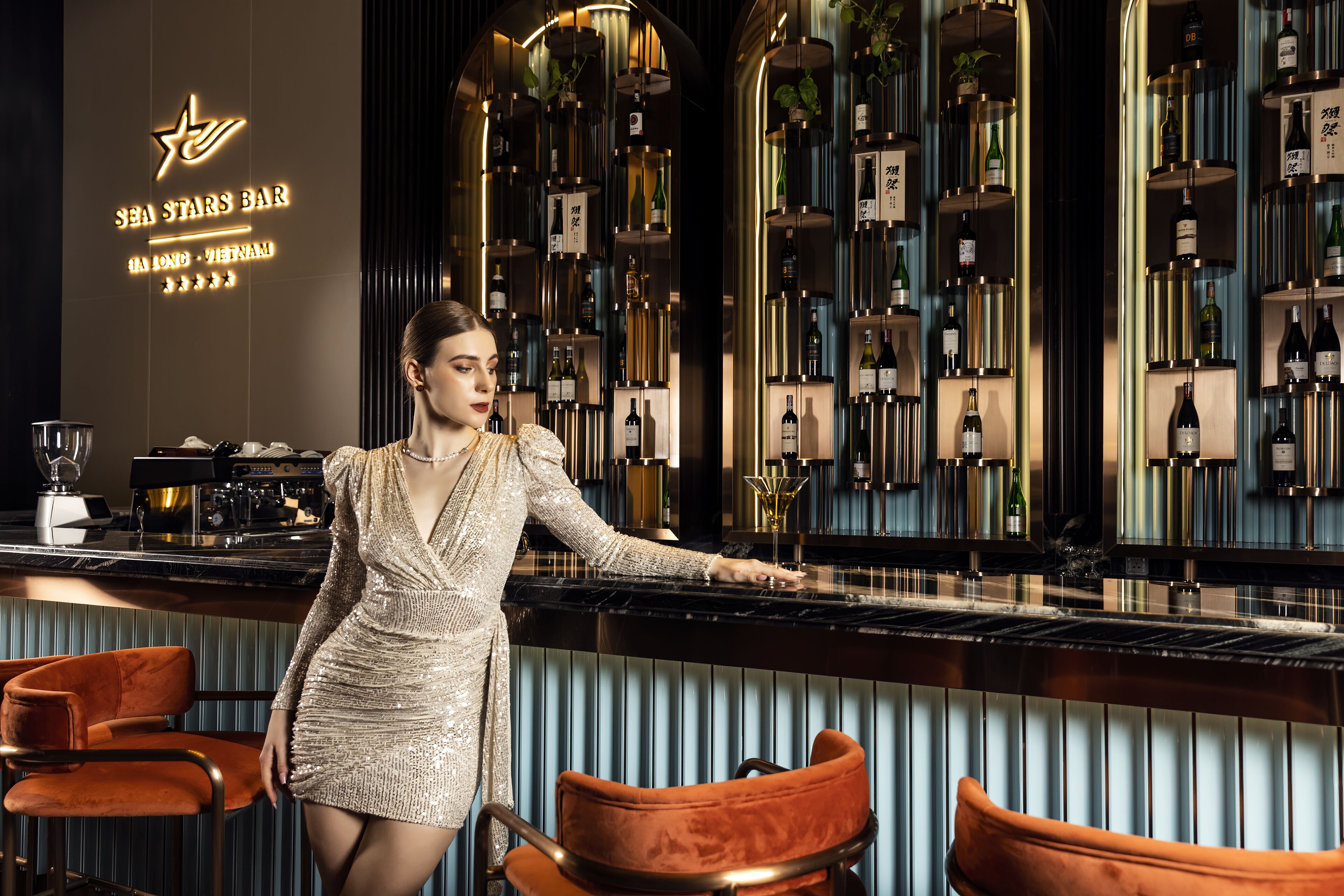 A bar area with a woman in a dress, bar stools, and a wall of wine bottles.