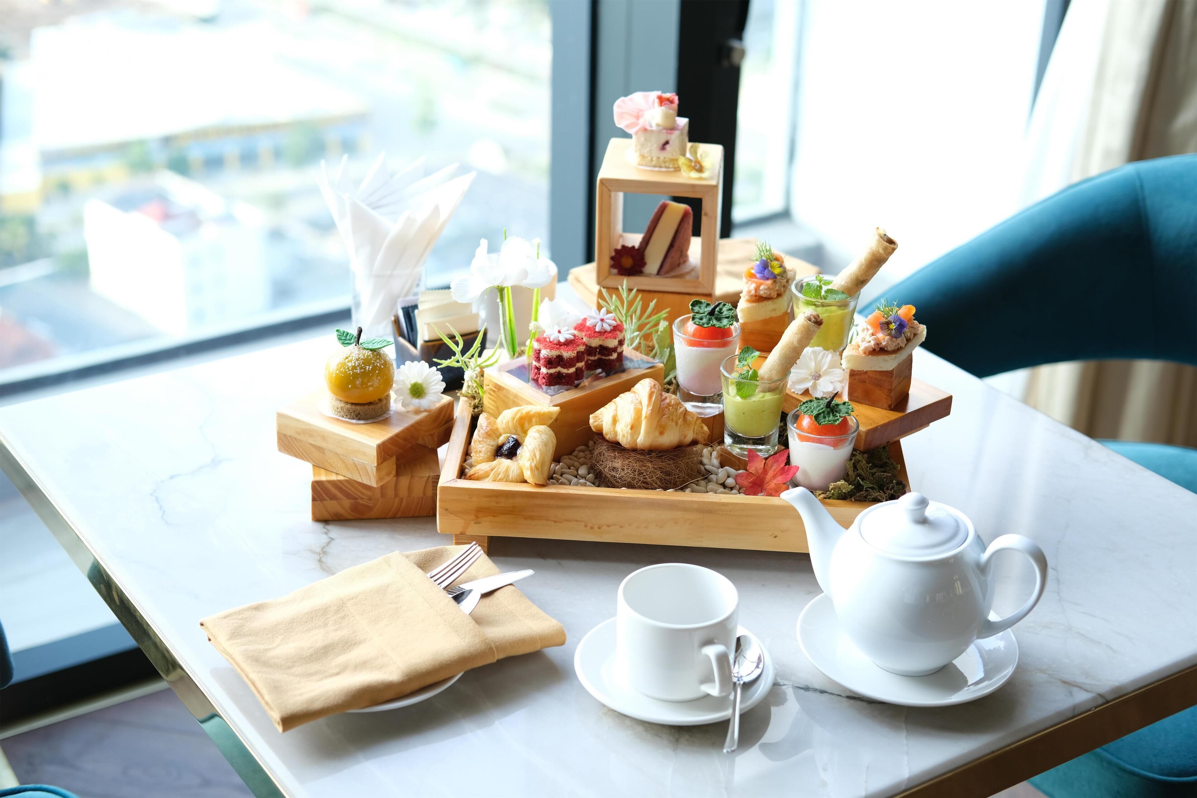 A breakfast setup with pastries, fruits, and a teapot on a table by a window.