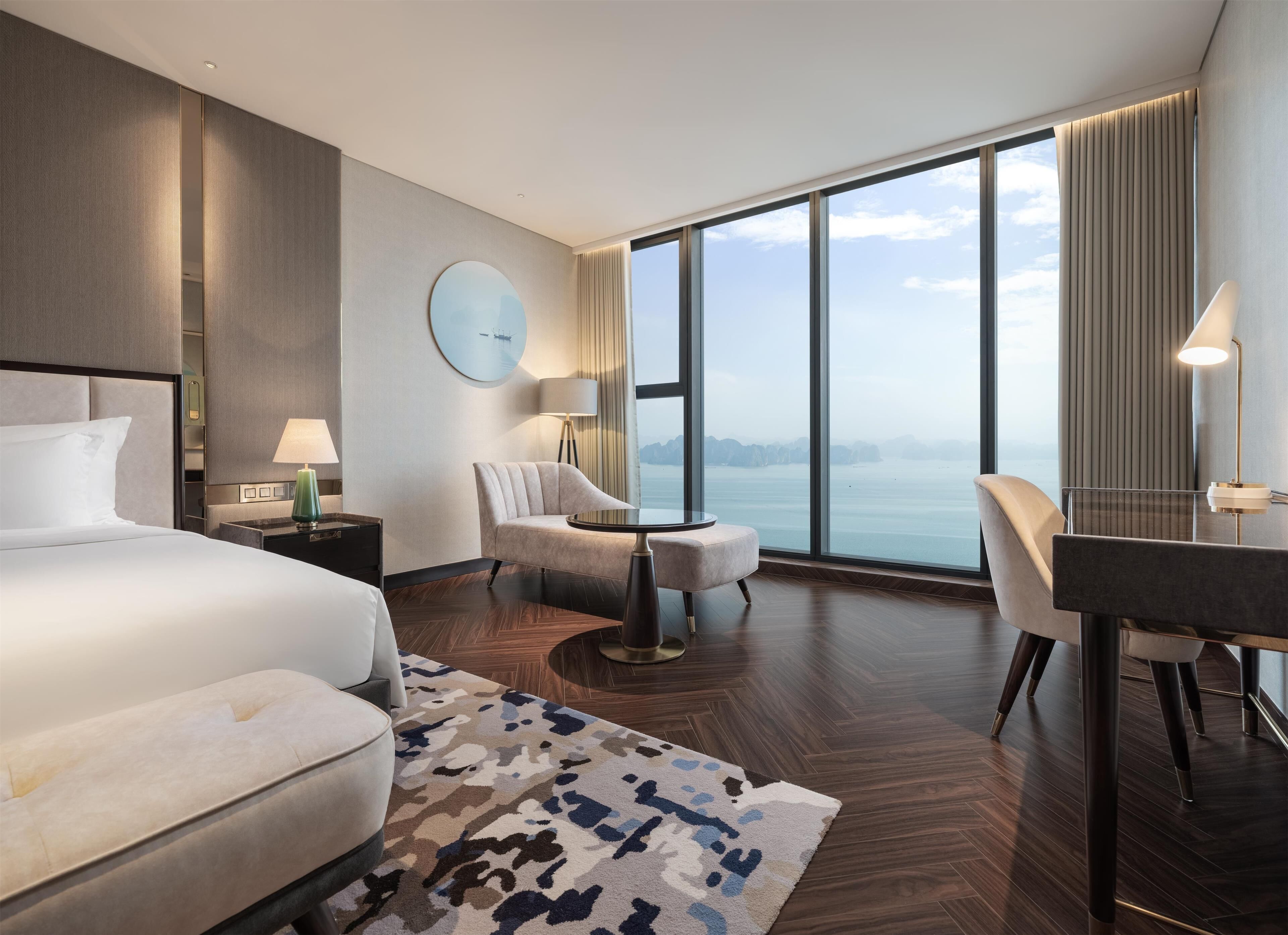 Habitación de hotel moderna con una cama grande, un escritorio, una silla y vista al mar a través de amplias ventanas.