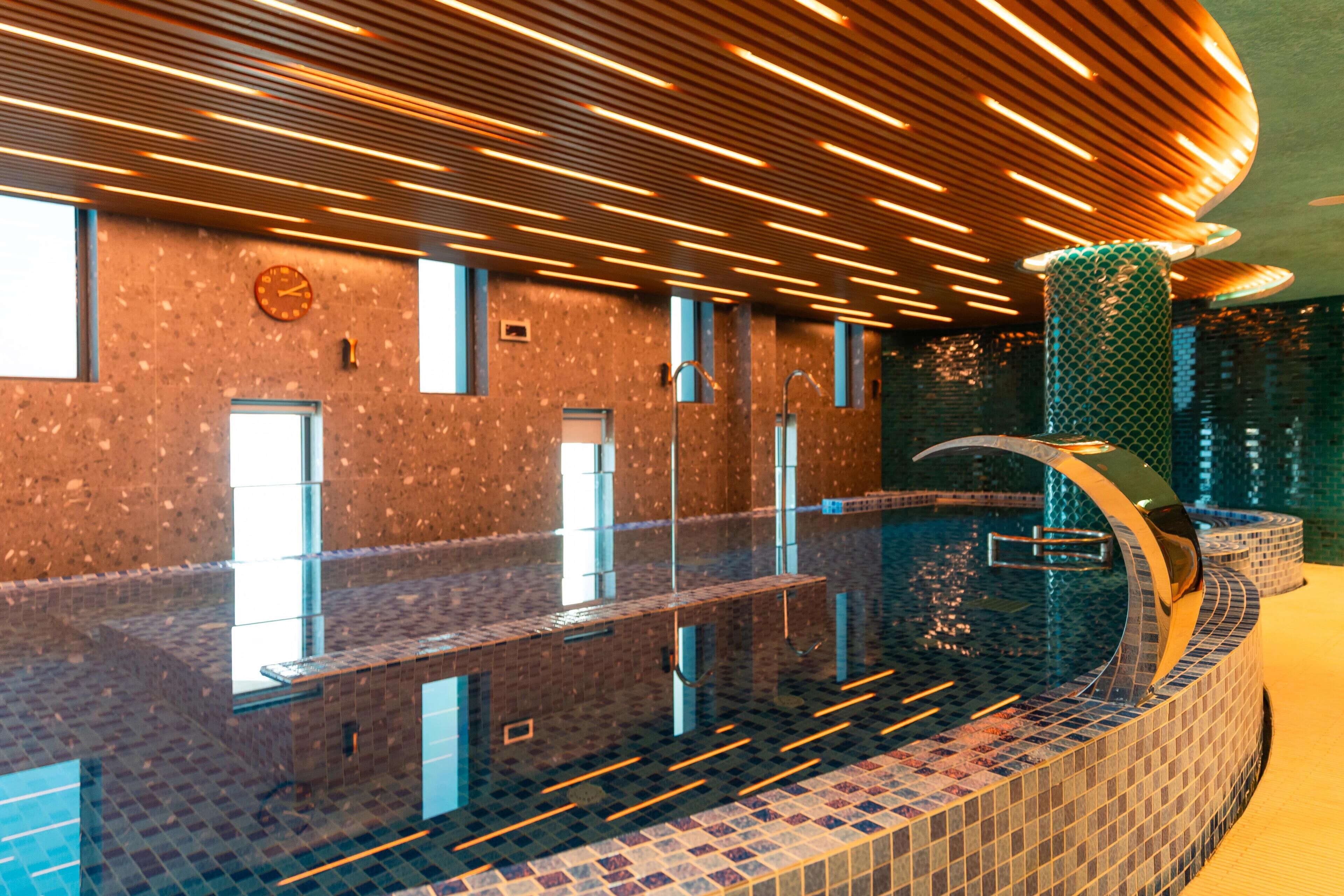 Une piscine intérieure moderne, dotée d’un sol carrelé et d’un bord incurvé. Des fenêtres s’étendent sur toute la longueur de la piscine, et le plafond est agrémenté de luminaires linéaires.