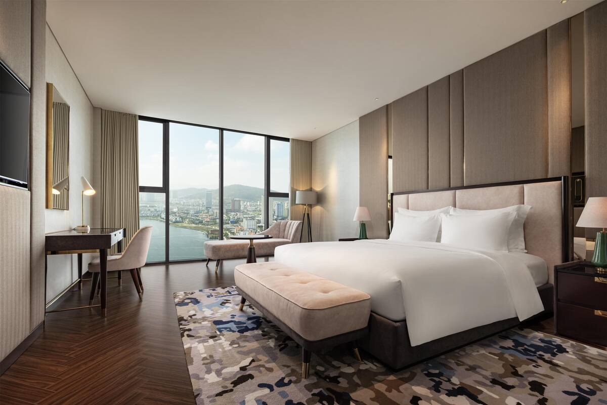 A modern hotel room with a large bed, a desk, a chair, and a view of the city.