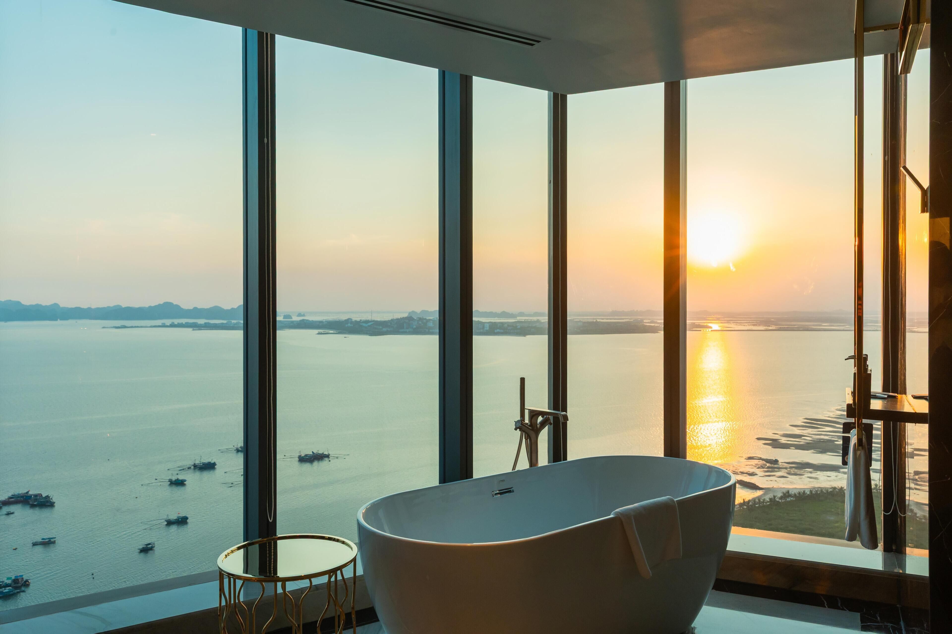 Une salle de bain moderne avec une baignoire sur pieds, de grandes fenêtres et une vue sur un plan d'eau au coucher du soleil.