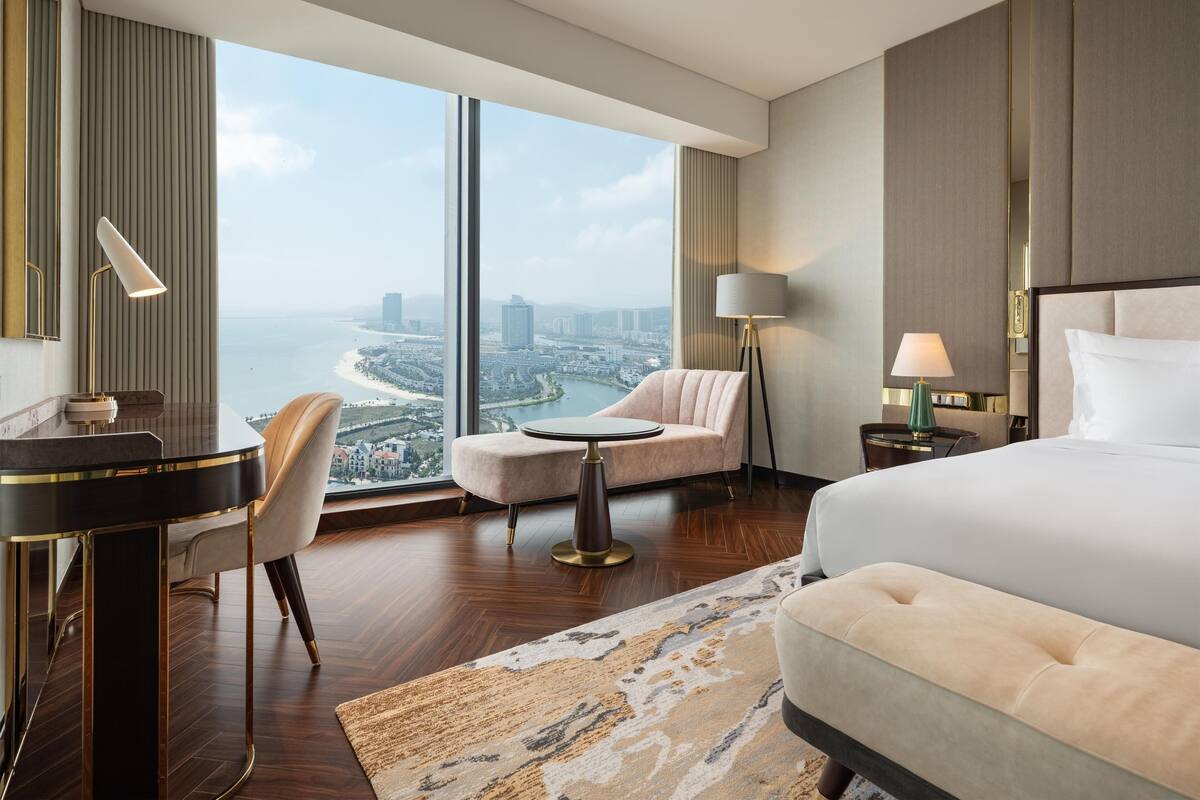 A modern hotel room with a large window, a bed, a desk, a chair, and a sofa.