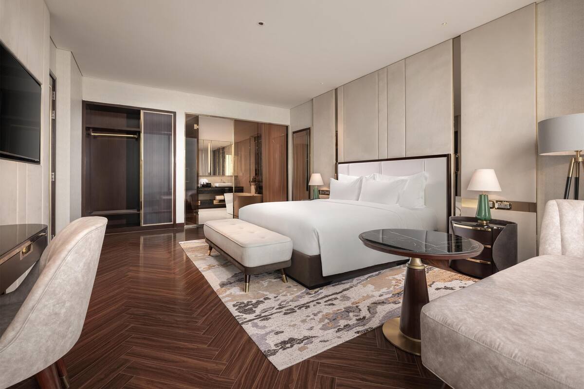 A modern hotel room with a large bed, a desk, a chair, a TV, and a bathroom visible through an open door.