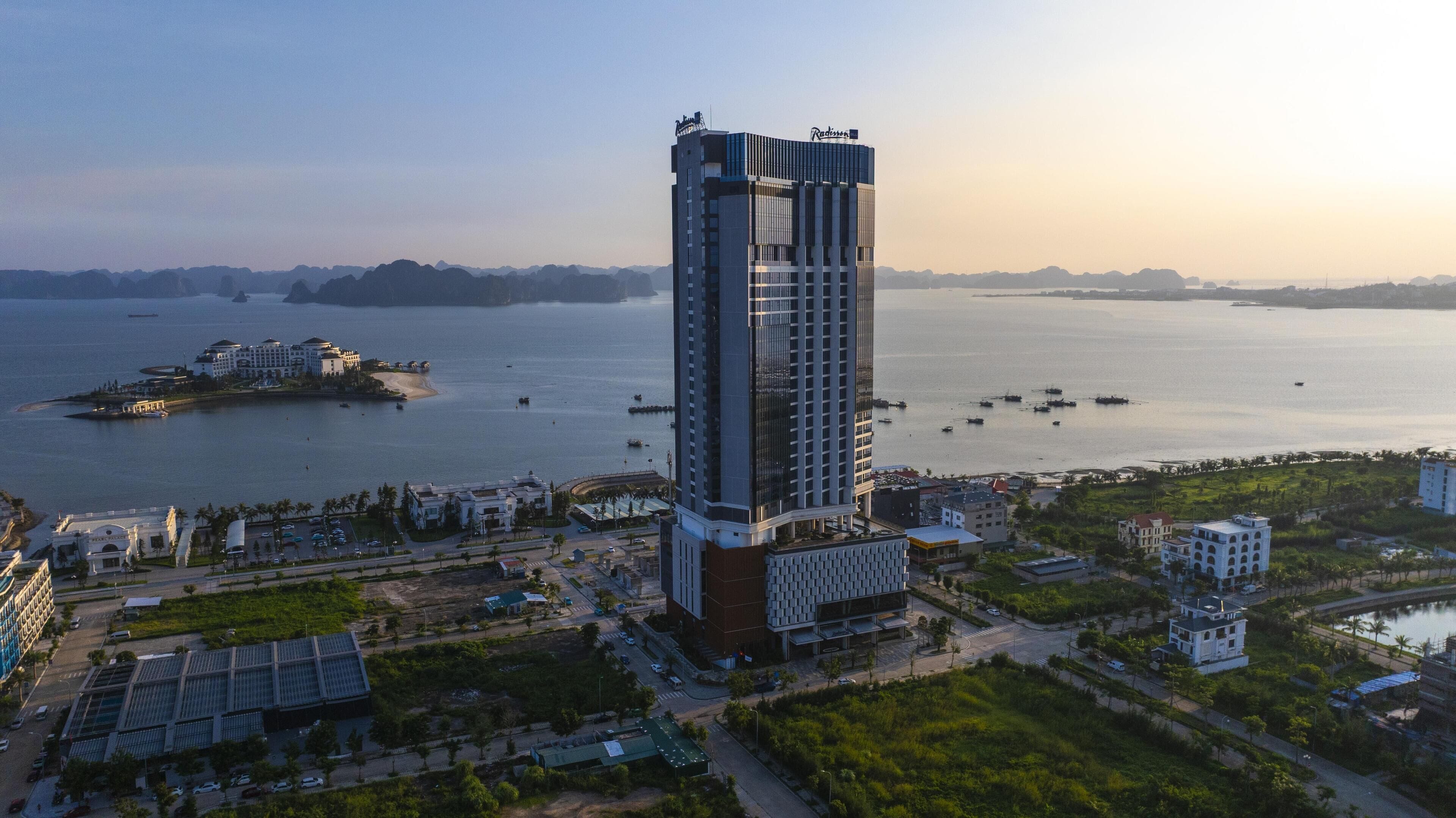 A high-rise hotel with a modern architectural design, situated near a body of water with boats and a distant island.