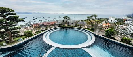A swimming pool with a curved design, surrounded by a fence and palm trees, overlooking a body of water with boats.