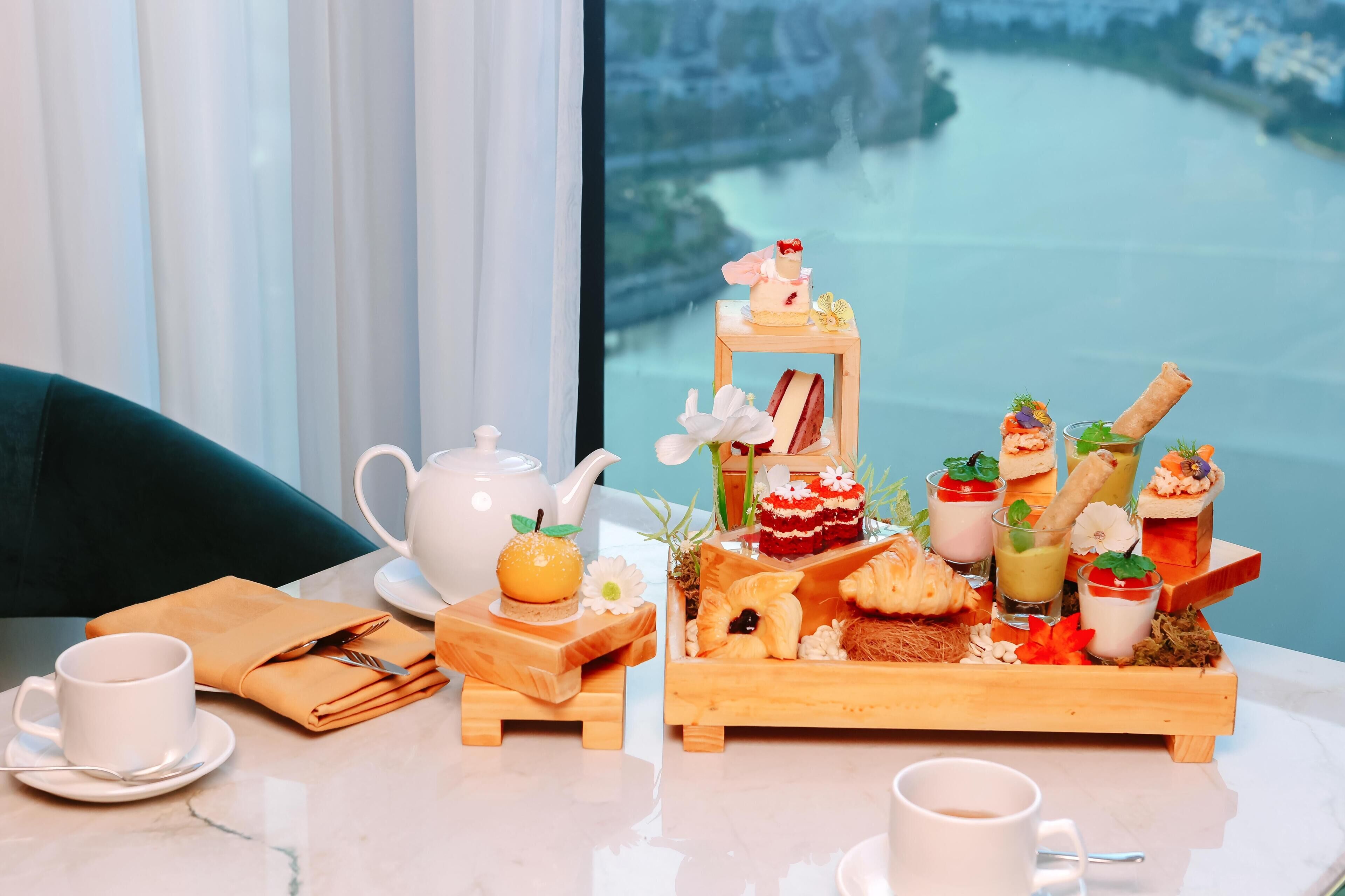 Une table dressée avec une théière, des tasses, une serviette, un plateau contenant diverses pâtisseries et desserts, et une vue pittoresque par la fenêtre.