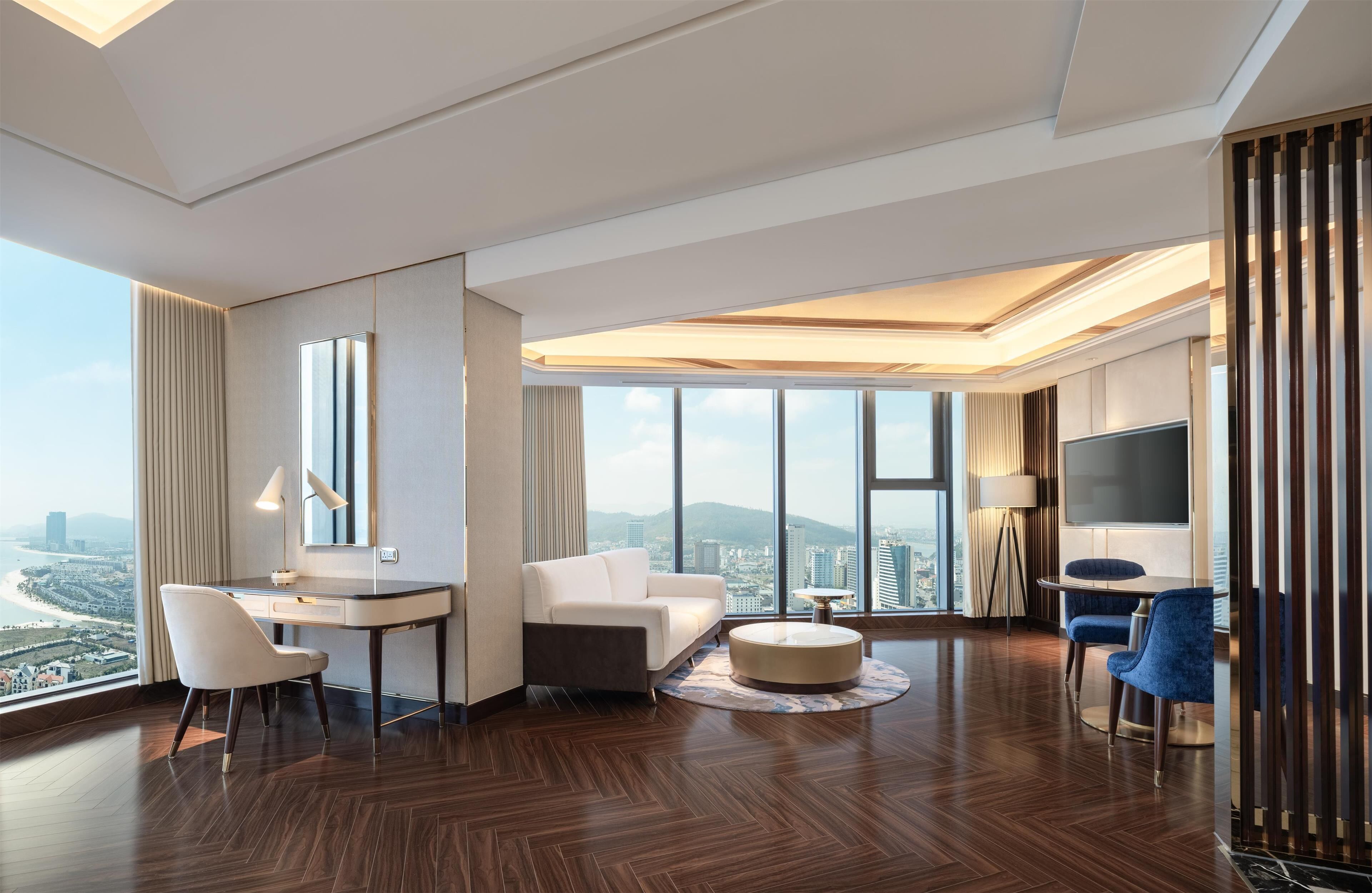 A modern hotel room with a large window, a desk, a sofa, a TV, and a dining area.