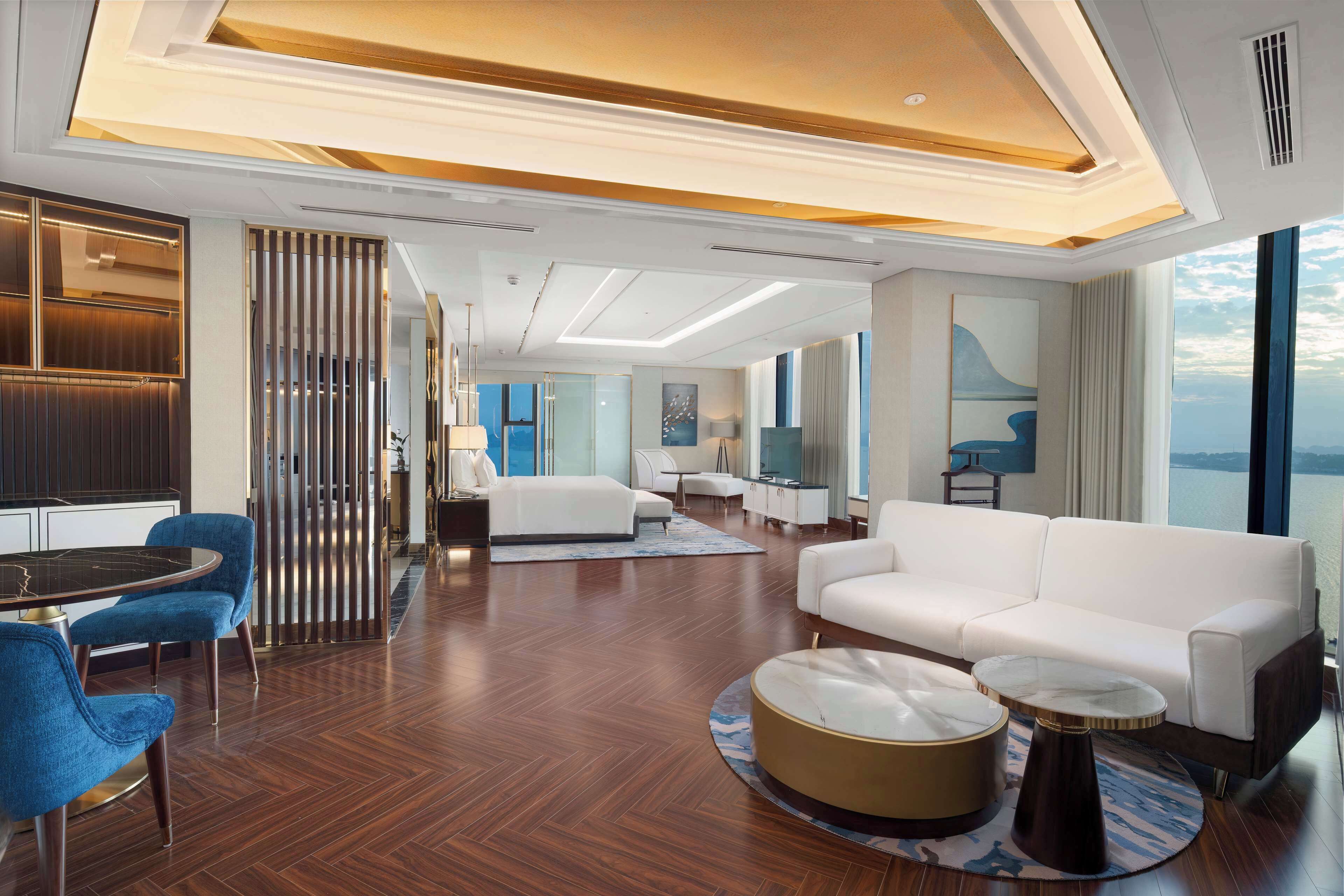 A modern hotel room with a wooden floor, a white sofa, a round coffee table, and a dining area with blue chairs.