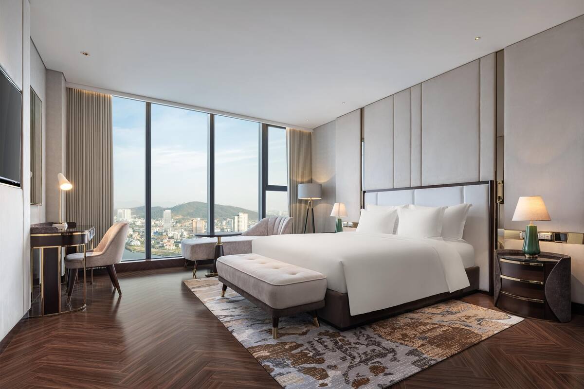 A modern hotel room with a large bed, a desk, a chair, and a view of the city.