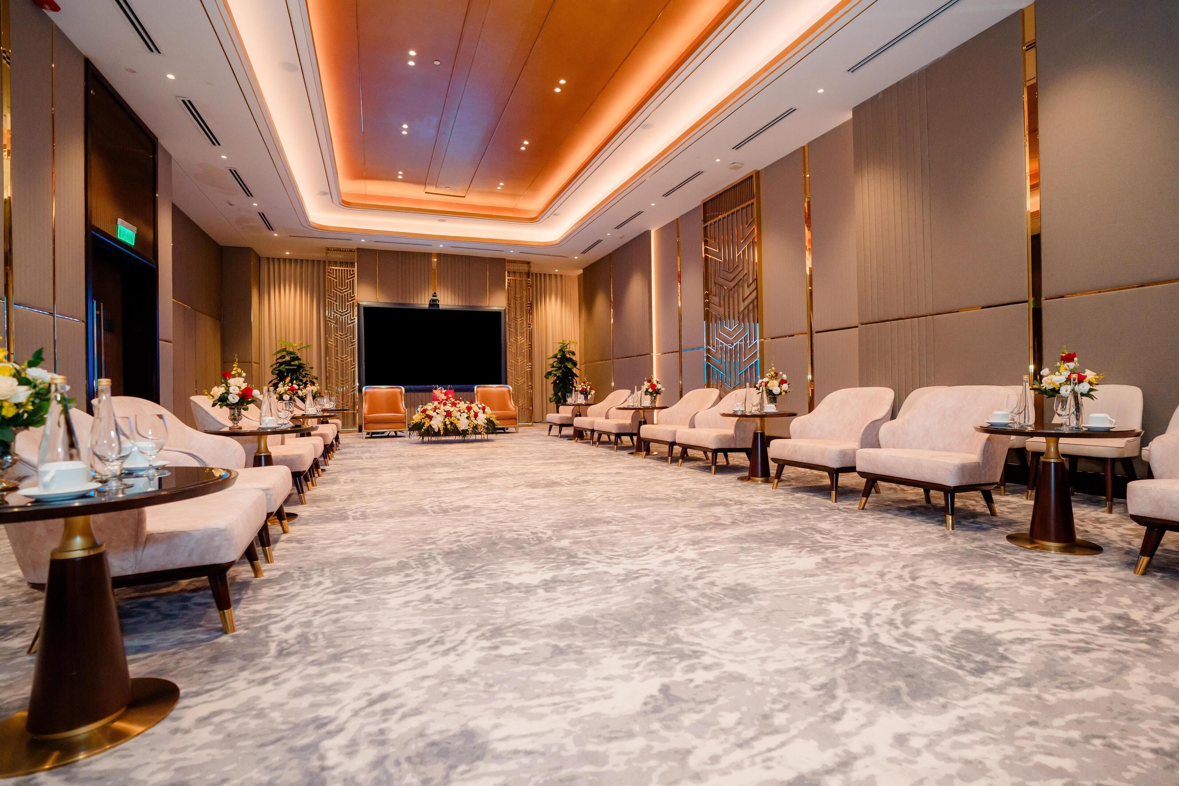 Une salle de conférence spacieuse, dotée d’un grand écran, de rangées de sièges et de tables ornées de compositions florales.