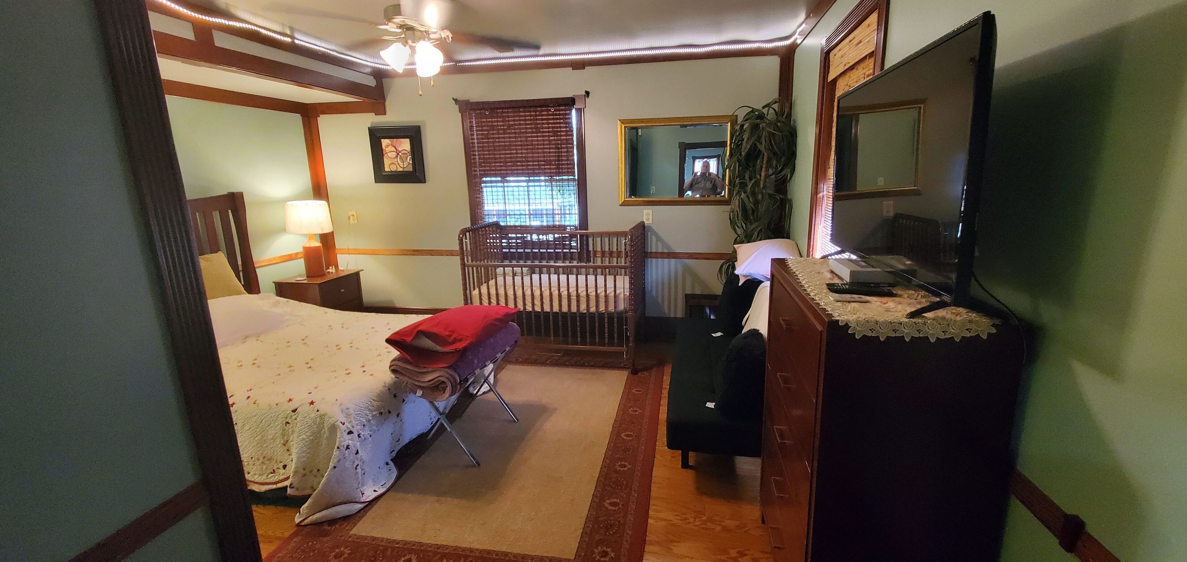 Ein Schlafzimmer mit einem Bett, einem Schreibtisch, einem Stuhl, einem Babybett, einem Spiegel und einem Fenster mit Jalousien.