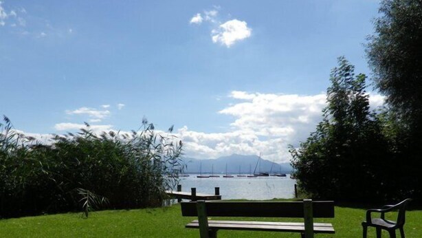 A park bench overlooking a body of water with boats, a mountain in the distance, and a clear sky. - Wohnung 4 a - 1. Etage, 30qm - Eckbalkon (Gstadt am Chiemsee)