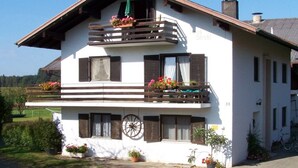 A two-story house with a balcony on the upper floor, a bicycle parked outside, and a number '38' on the side. - Wohnung 4 a - 1. Etage, 30qm - Eckbalkon (Gstadt am Chiemsee)