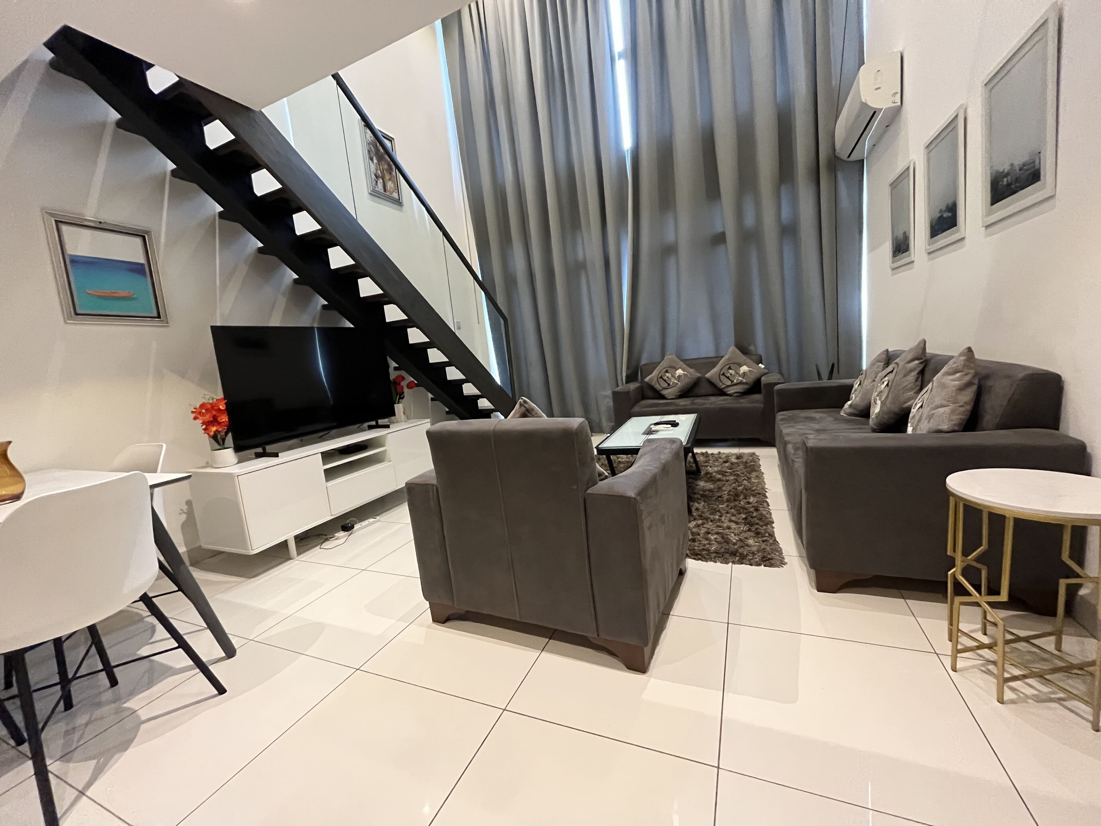 A modern living room with a staircase, grey sofas, a flat-screen TV, and a dining area.
