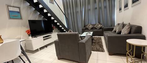 A modern living room with a staircase, grey sofas, a flat-screen TV, and a dining area.