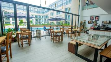 Un comedor interior moderno con mesas y sillas de madera, ventanas amplias y vistas a una piscina y edificios.
