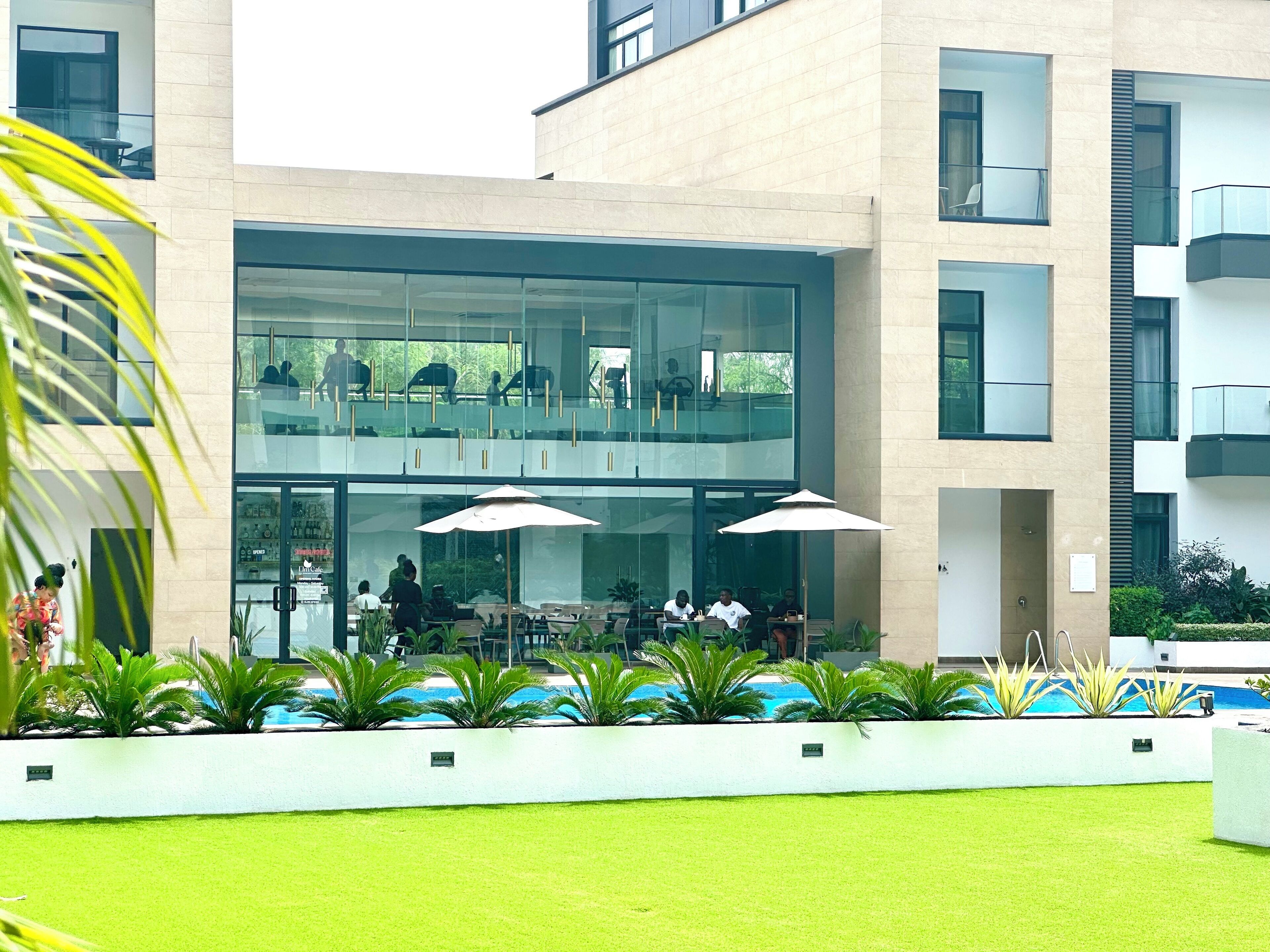 A modern building with a glass facade, outdoor seating area with umbrellas, and a pool.