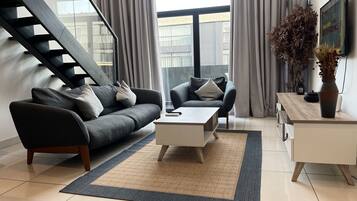 A modern living room with a grey sofa, a coffee table, and a TV on a wooden cabinet. A staircase is visible in the background.