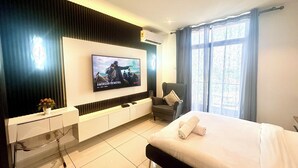 A modern hotel room with a large flat-screen TV on the wall, a bed with white linens, a seating area with a chair, and a small table with a vase of flowers. - Unique Homes by Embassy Gardens (Accra)