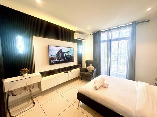 A modern hotel room with a large flat-screen TV on the wall, a bed with white linens, a seating area with a chair, and a small table with a vase of flowers. - Unique Homes by Embassy Gardens (Accra)