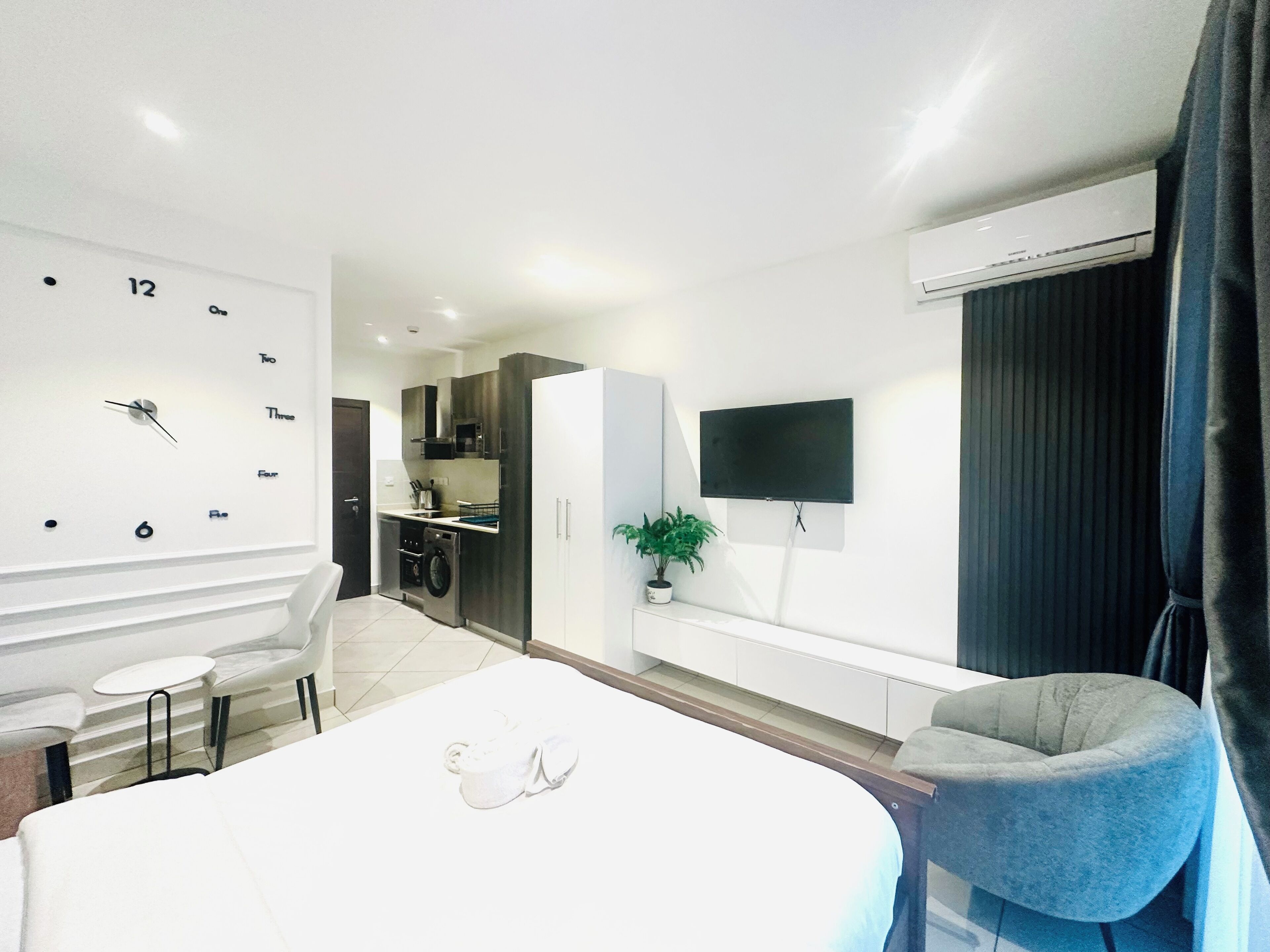 Et moderne hotelværelse med seng, siddeområde, lille tekøkken, fjernsyn ophængt på væggen og et klimaanlæg.
