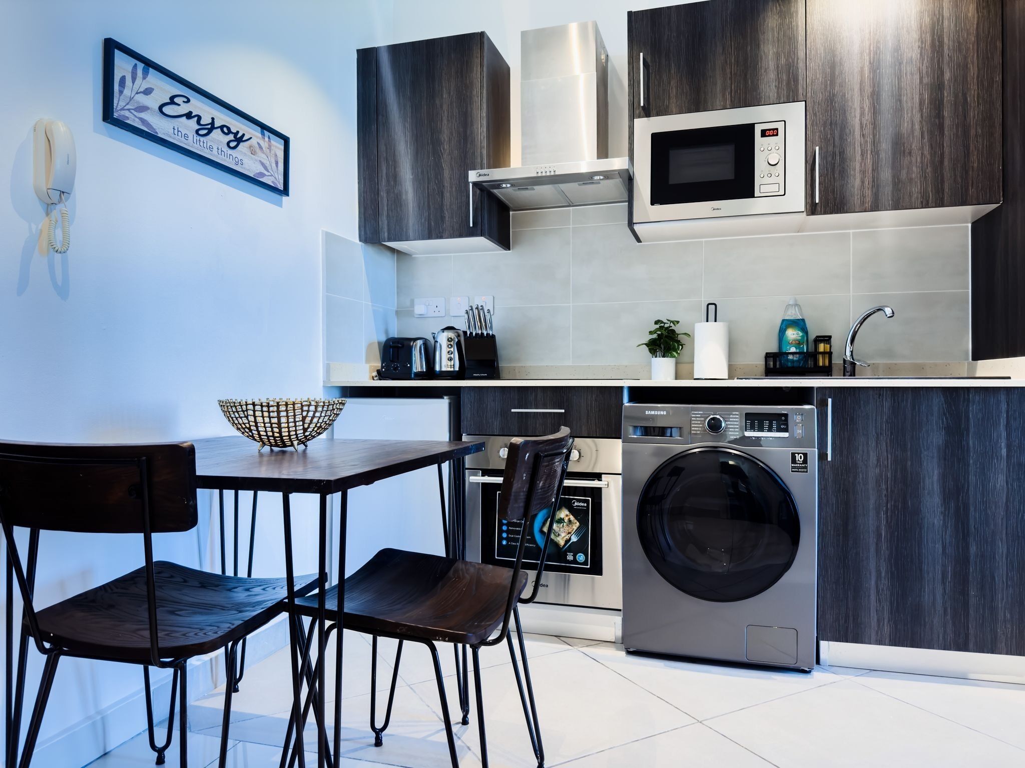 A modern kitchen with dark wood cabinets, a microwave, a washing machine, and a dining area with two chairs.
