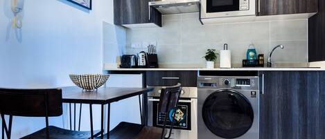 A modern kitchen with dark wood cabinets, a microwave, a washing machine, and a dining area with two chairs.