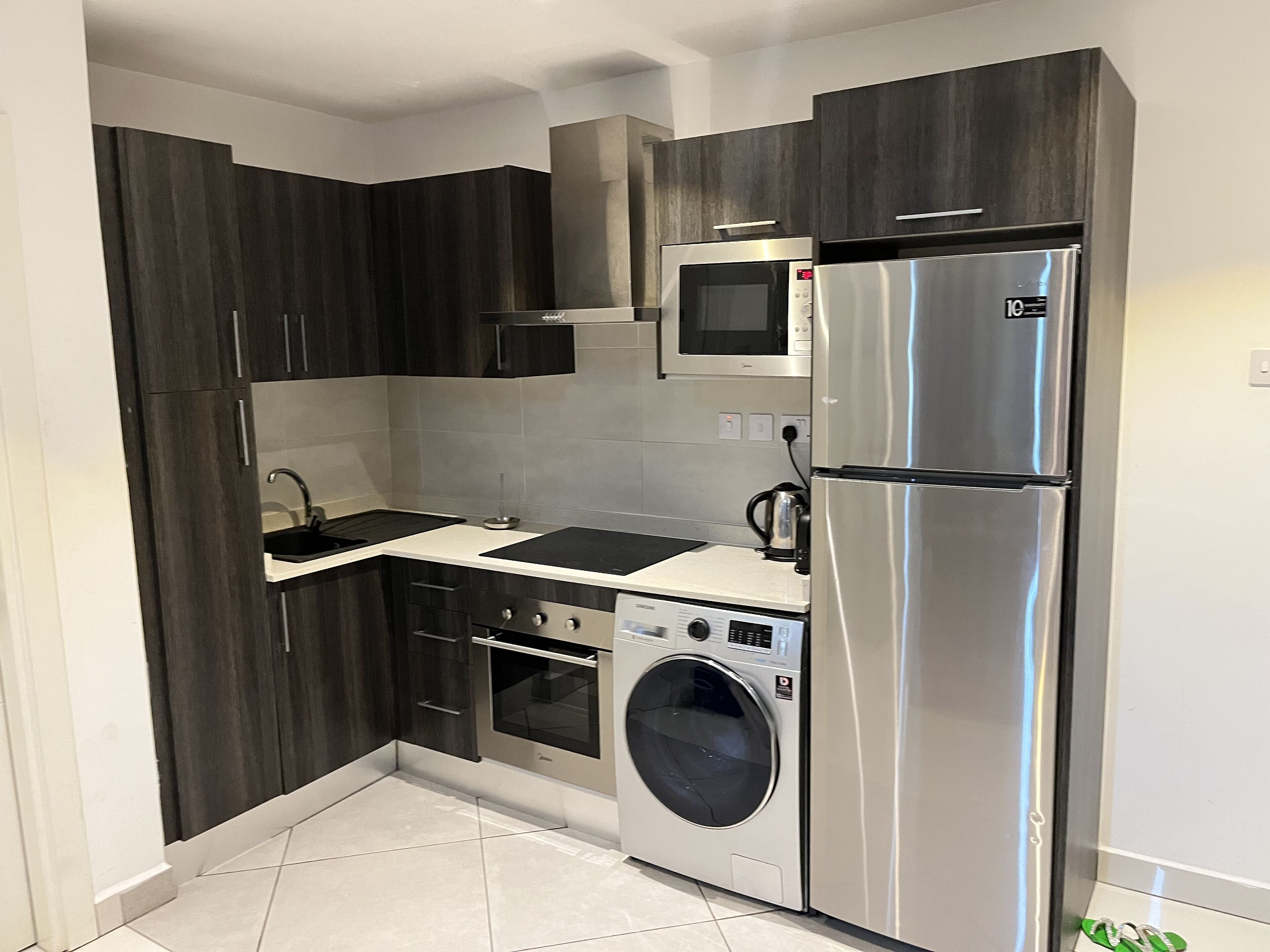 Une cuisine compacte avec des meubles en bois foncé, un réfrigérateur en acier inoxydable, un four encastré, un four à micro-ondes et un lave-linge.