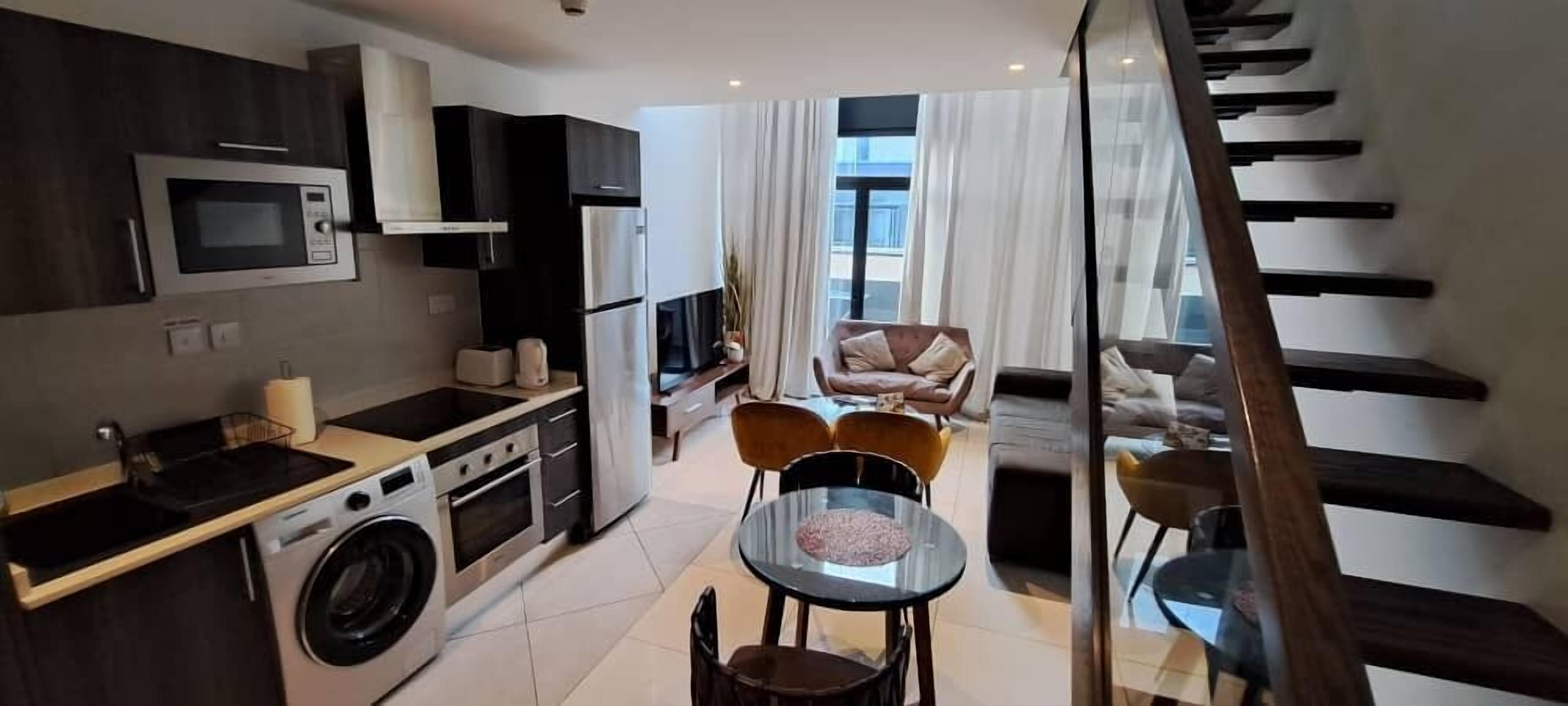 A compact kitchen with modern appliances, a dining area with a glass table, and a staircase leading to an upper level.