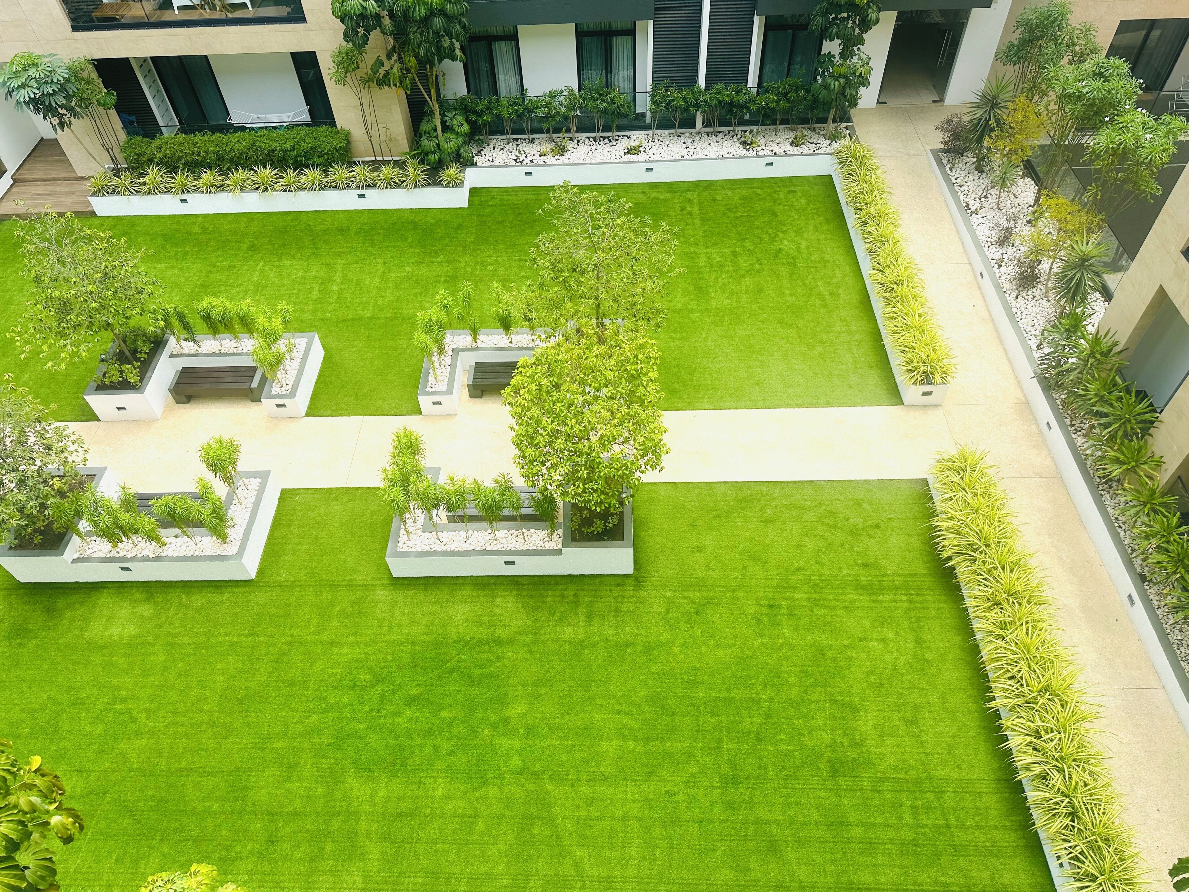 A courtyard with a central lawn, surrounded by planters with various plants and trees, and bordered by a building with large windows and a balcony.