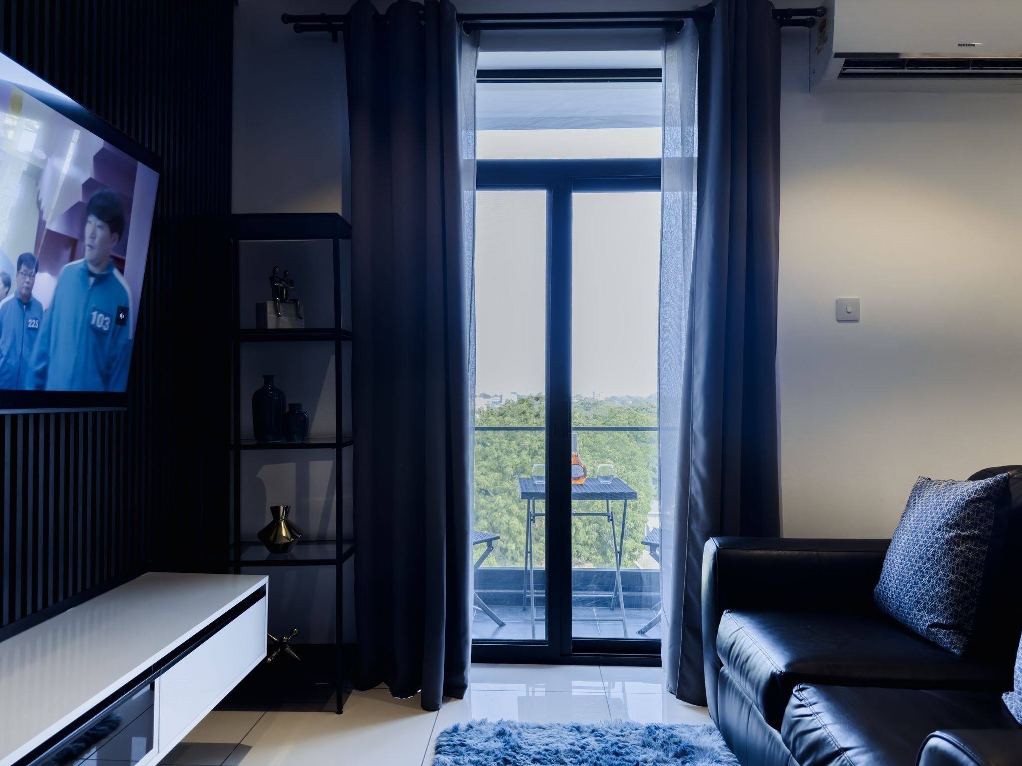 Ein modernes Wohnzimmer mit Fernseher, schwarzer Ledersofa und einem Balkon mit Tisch und Stühlen.