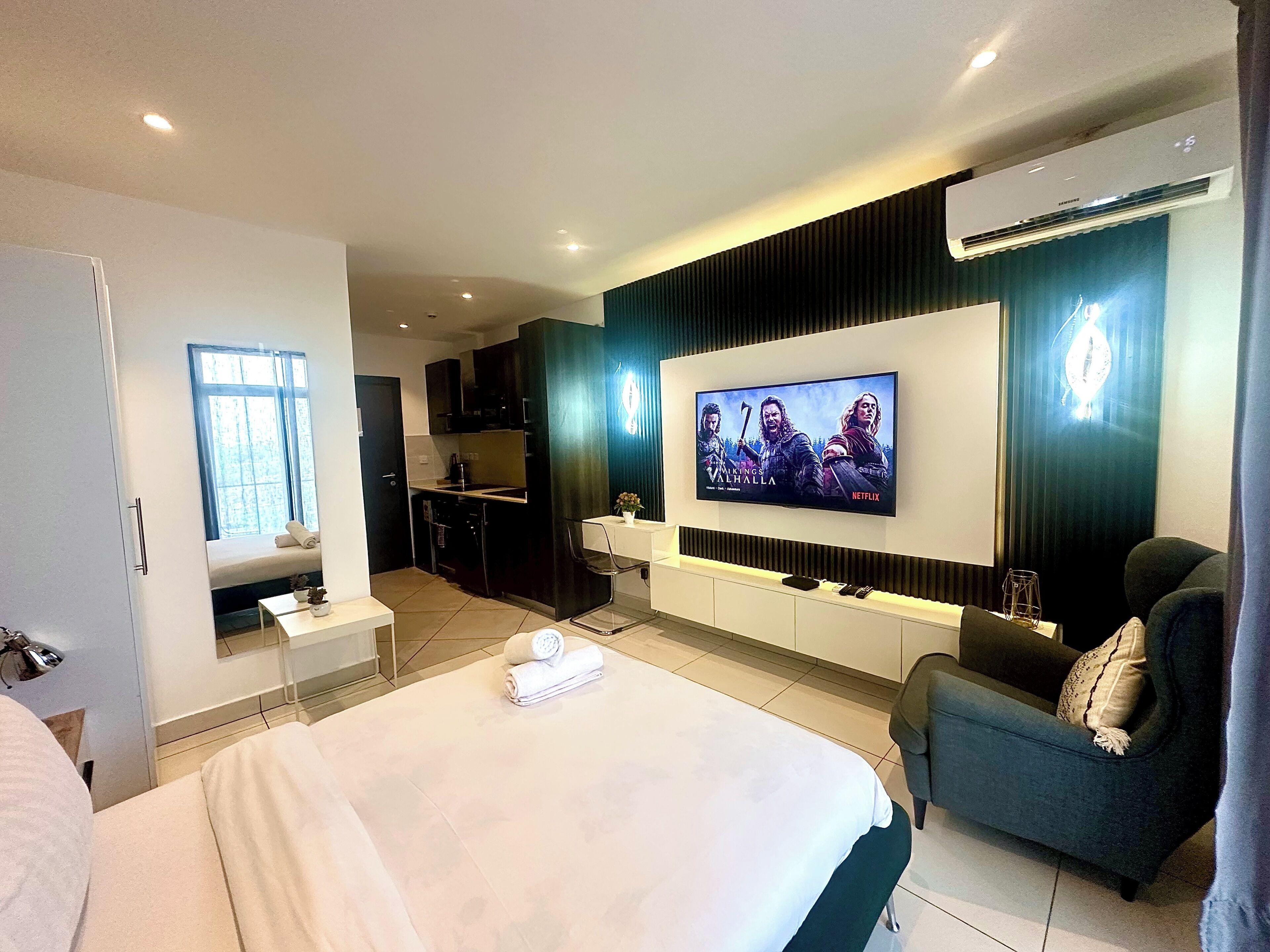 Ein modernes Hotelzimmer mit einem großen Bett, einem an der Wand befestigten Fernseher, einer kleinen Küchenzeile und einer Sitzecke mit Sofa und Sessel.