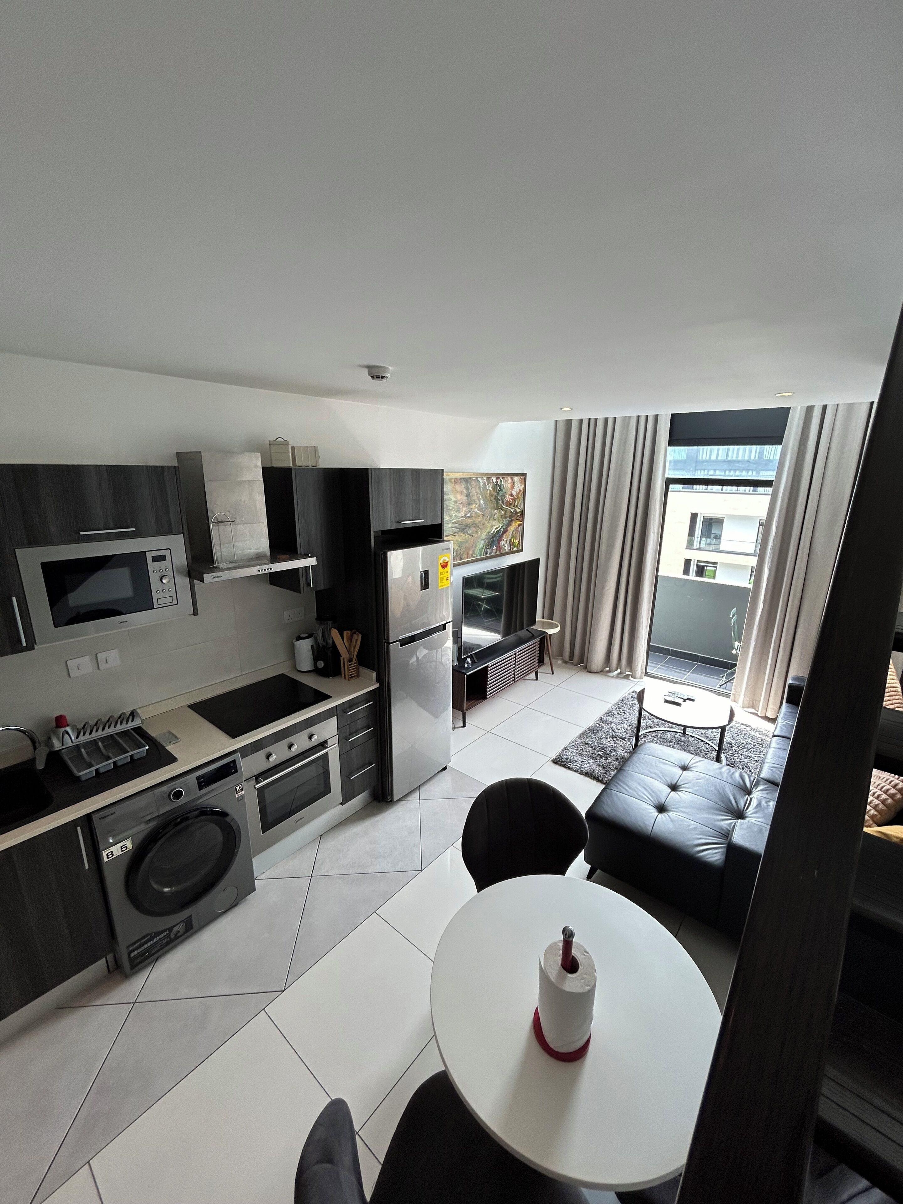 A modern kitchen with a microwave, stove, and refrigerator. There is a dining area with a table and chairs, and a view of the outside through large windows.