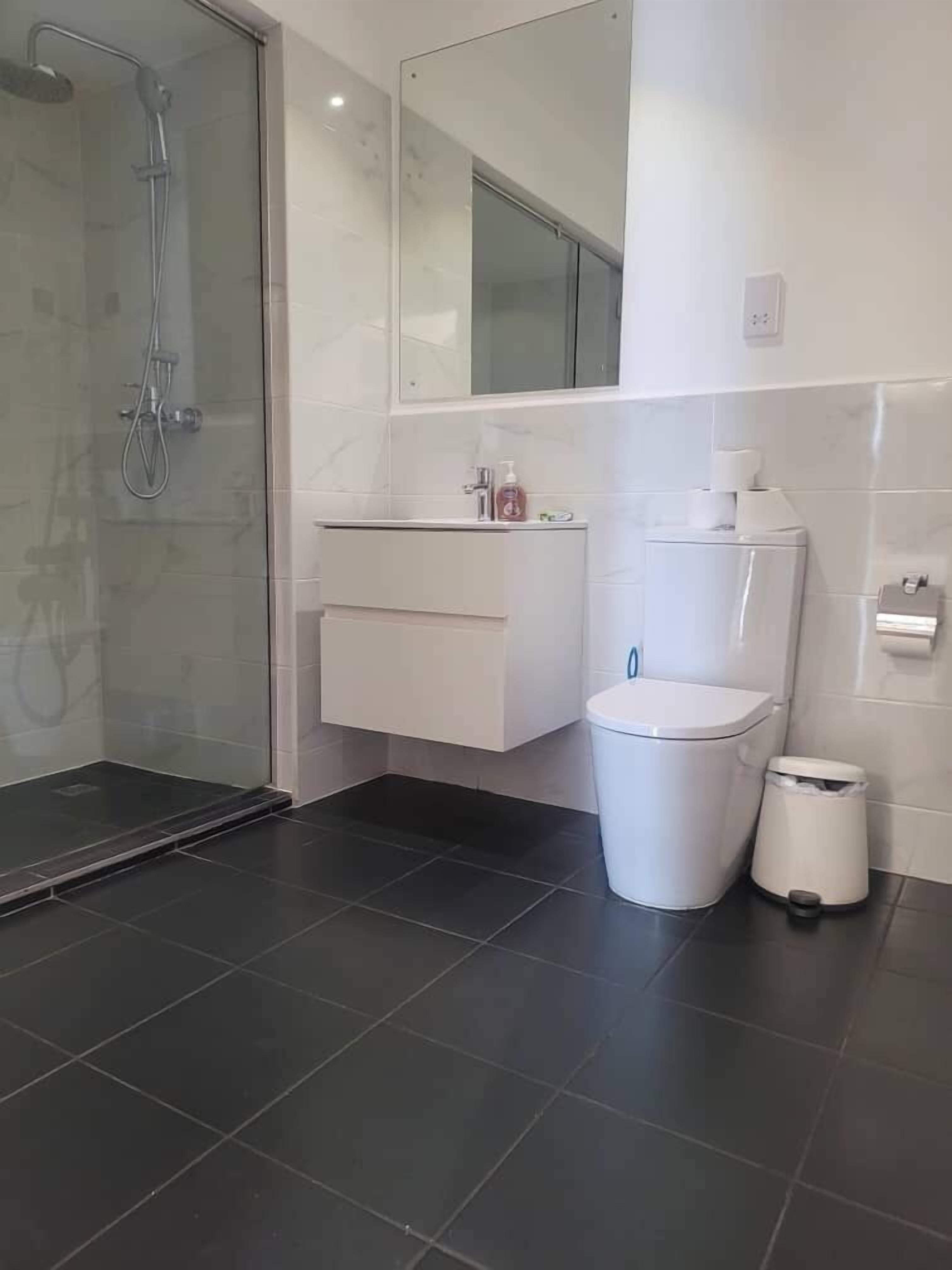 A modern bathroom with a glass-enclosed shower, a white vanity, a toilet, and a trash bin.