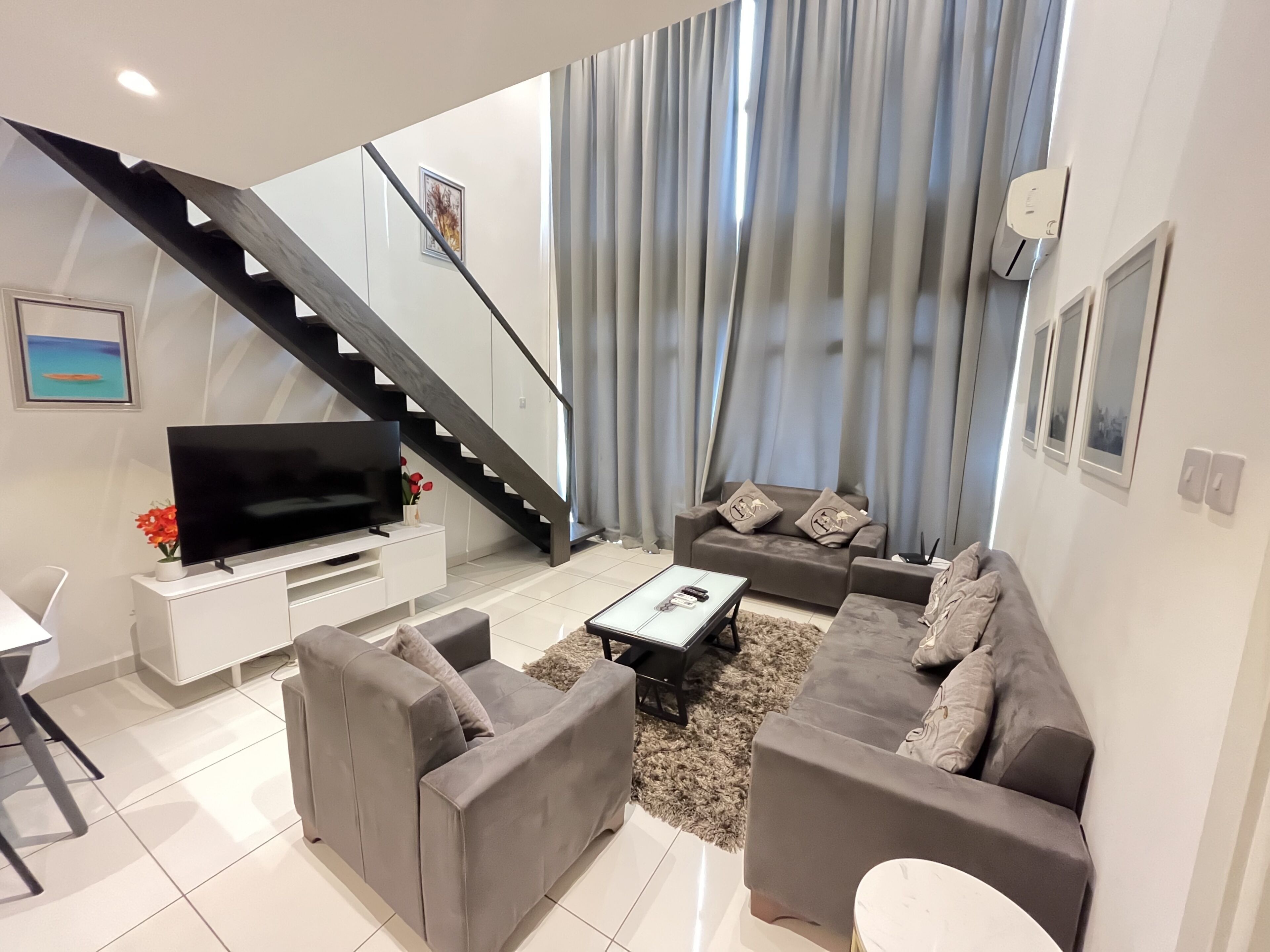 Una sala de estar moderna con escalera, sofás grises, un televisor de pantalla plana y una mesa de centro blanca.