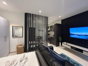 Una sala de estar moderna con un sofá negro, un televisor y una cocina al fondo.