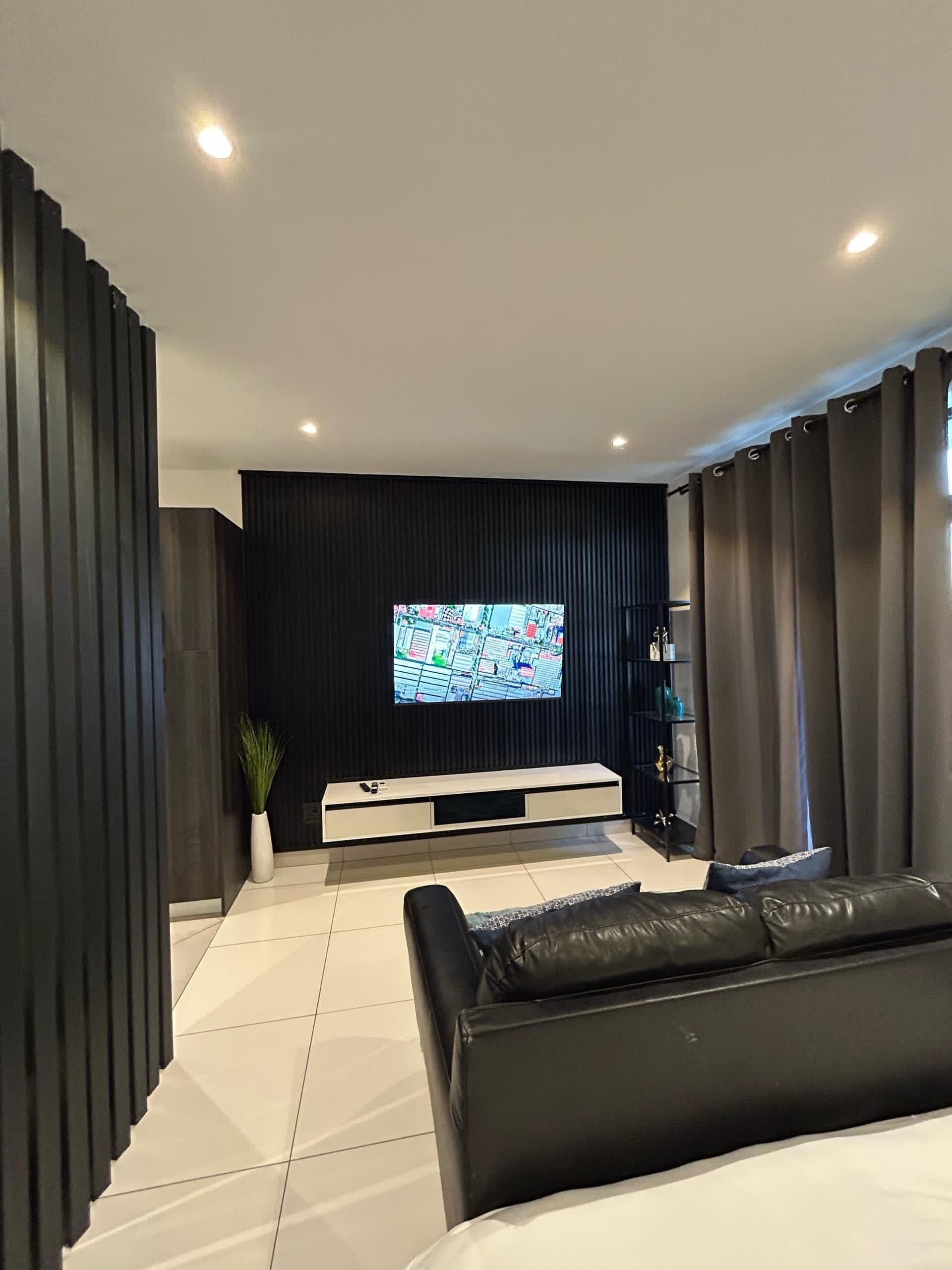 A modern living room with a black entertainment unit, a flat-screen TV mounted on the wall, a black leather sofa, and a potted plant.