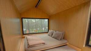 Una habitación pequeña y de madera con una cama, un ventanal y vista a árboles.