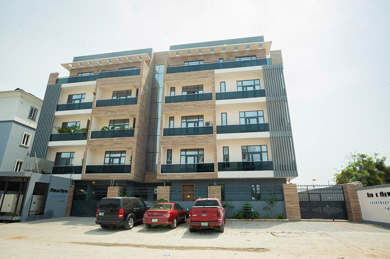 A multi-story residential building with balconies, a gated entrance, and parked cars in front.