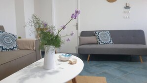 Ein Wohnzimmer mit einem weißen runden Tisch, einem grauen Sofa mit gemusterten Kissen und einem Blumentopf mit violetten Blüten.