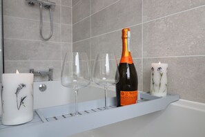 A bathroom with a bathtub, a shower, and a shelf with a bottle of wine, glasses, and candles.