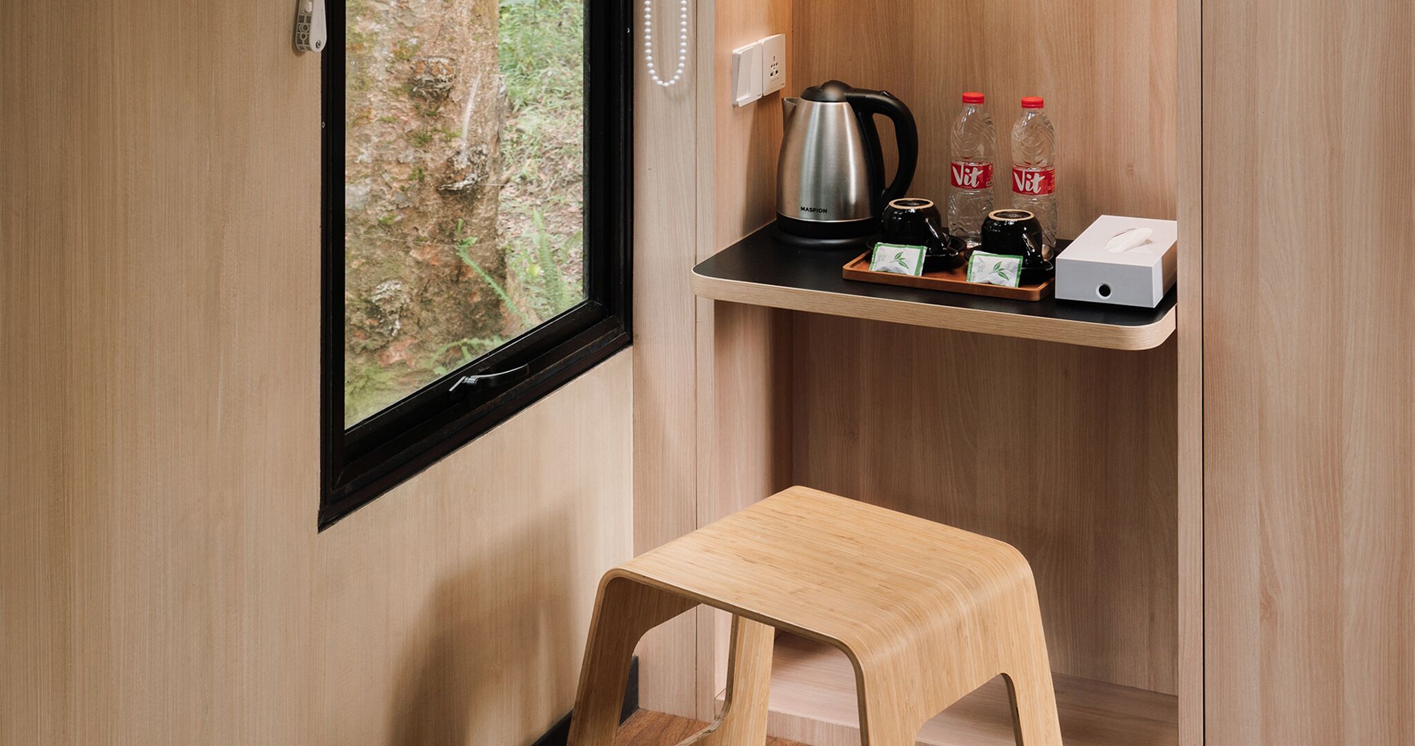 A small wooden stool, a window with a view of a forest, a shelf with a kettle, two bottles, and a box.