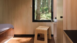 Una habitación pequeña con una cama, un taburete, un ventanal con vista a un bosque y una encimera de madera.