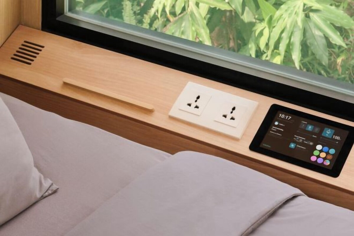 Una habitación moderna con ventana, una mesita de noche con una tableta mostrando información sobre la habitación y un enchufe.