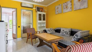 A bright yellow room with a wooden dining table, striped chairs, a sofa with patterned cushions, and a white cabinet.