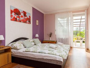 A bedroom with a bed, a nightstand, a lamp, a small table with a vase, and a view of the outside through a window.