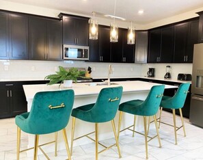 A modern kitchen with dark cabinets, a white island, and four teal chairs with gold legs.